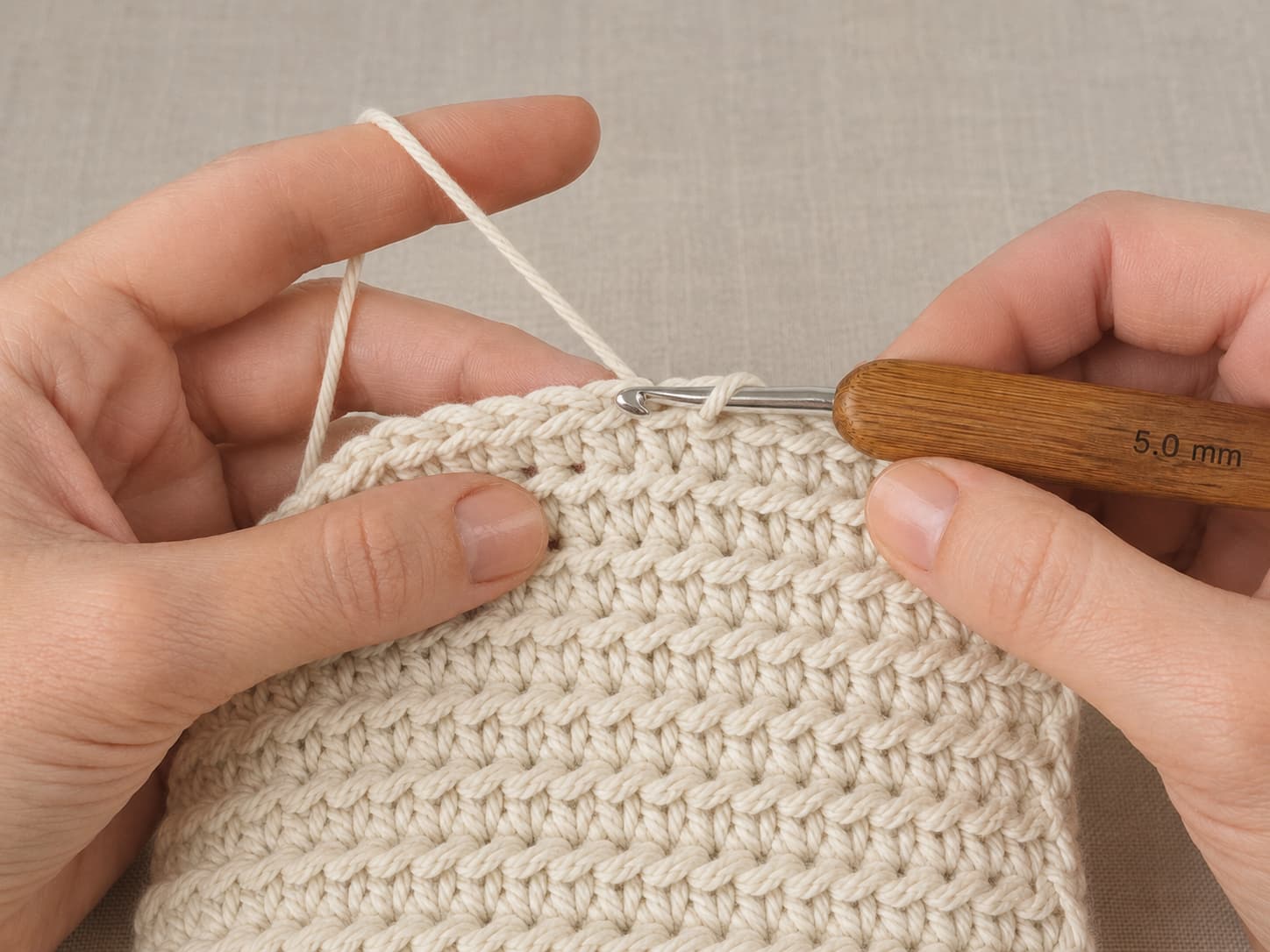Hands crocheting a cream cotton dishcloth with the hook inserted under one back loop above finished ridged rows.