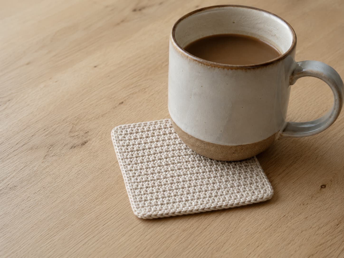 Finished square cotton single-crochet coaster beside a ceramic mug on a light wooden table.