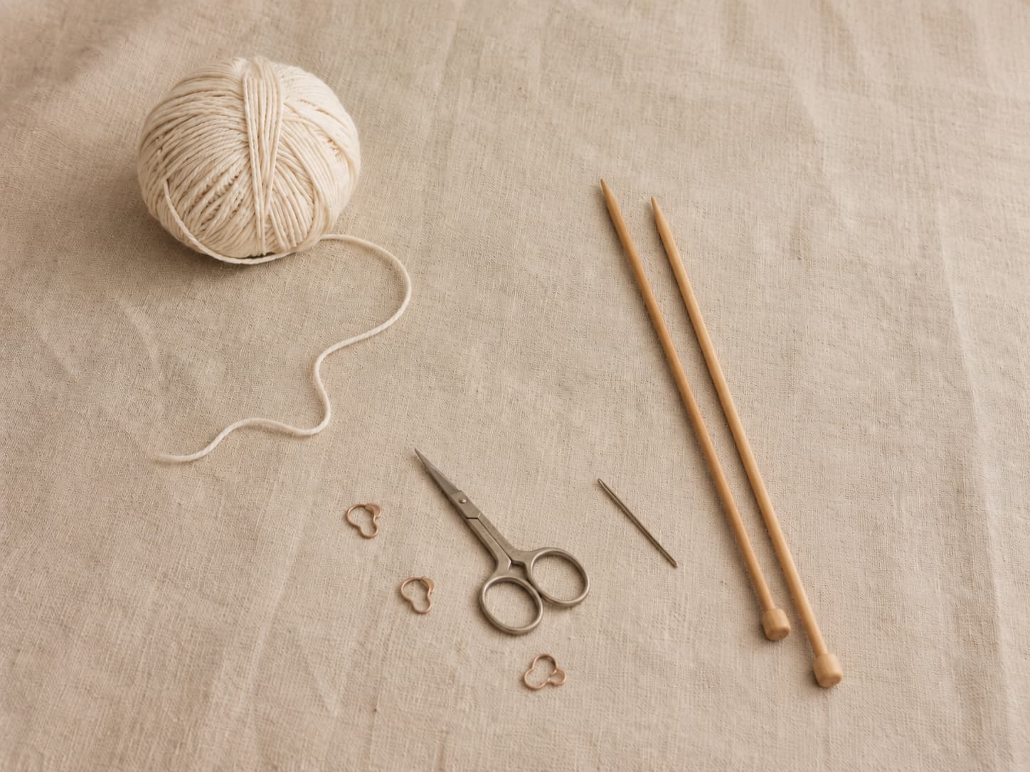 Wooden knitting needles, cream cotton yarn, small scissors, a tapestry needle, and stitch markers arranged on natural linen.