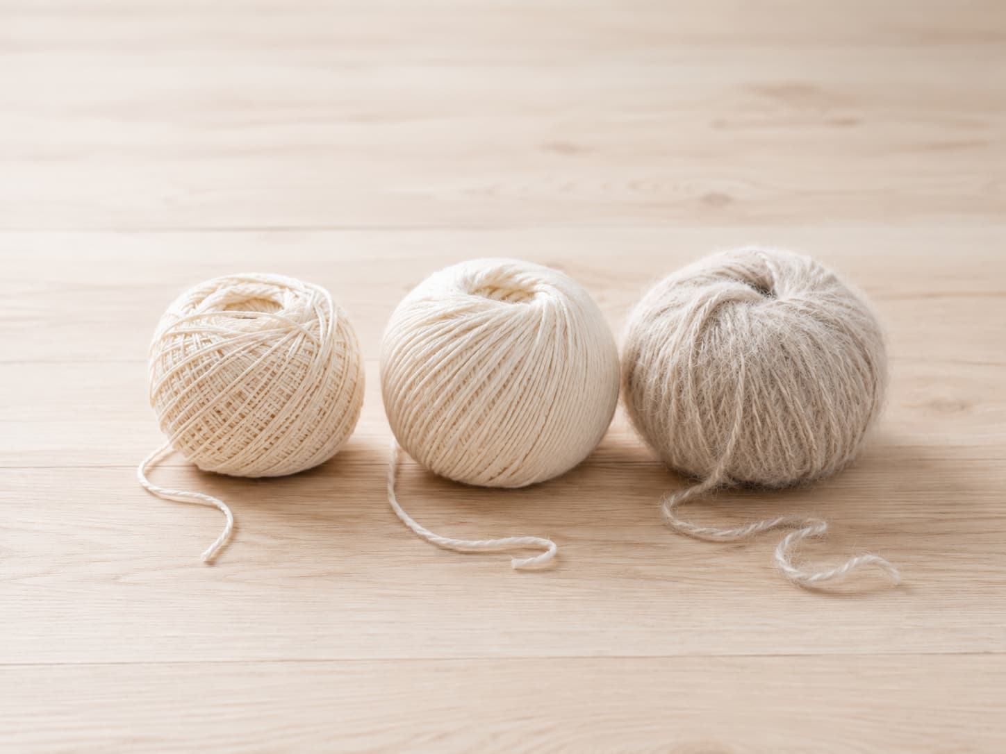 Cream cotton, acrylic, and wool yarn balls with loose strands arranged side by side on a light wooden table.