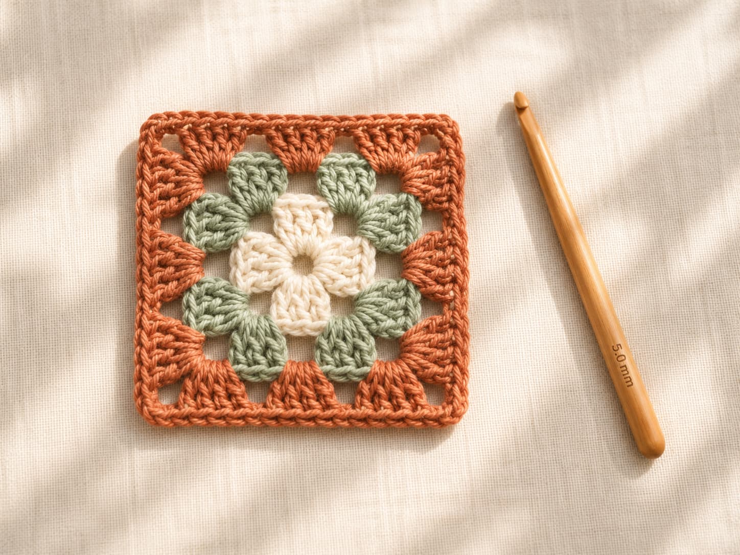 Classic three-round granny square with a cream center, sage middle round, and terracotta outer round beside a wooden crochet hook.