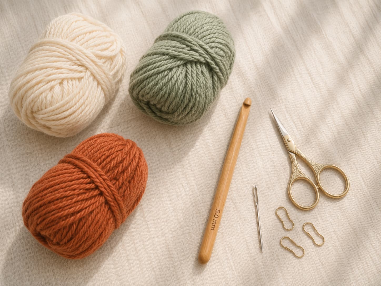 Cream, sage, and terracotta worsted yarn with a 5 mm wooden crochet hook, scissors, tapestry needle, and stitch markers on linen.