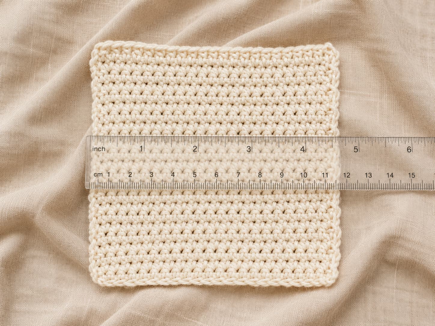Cream cotton crochet gauge swatch measured across the center with a clear ruler on natural linen.