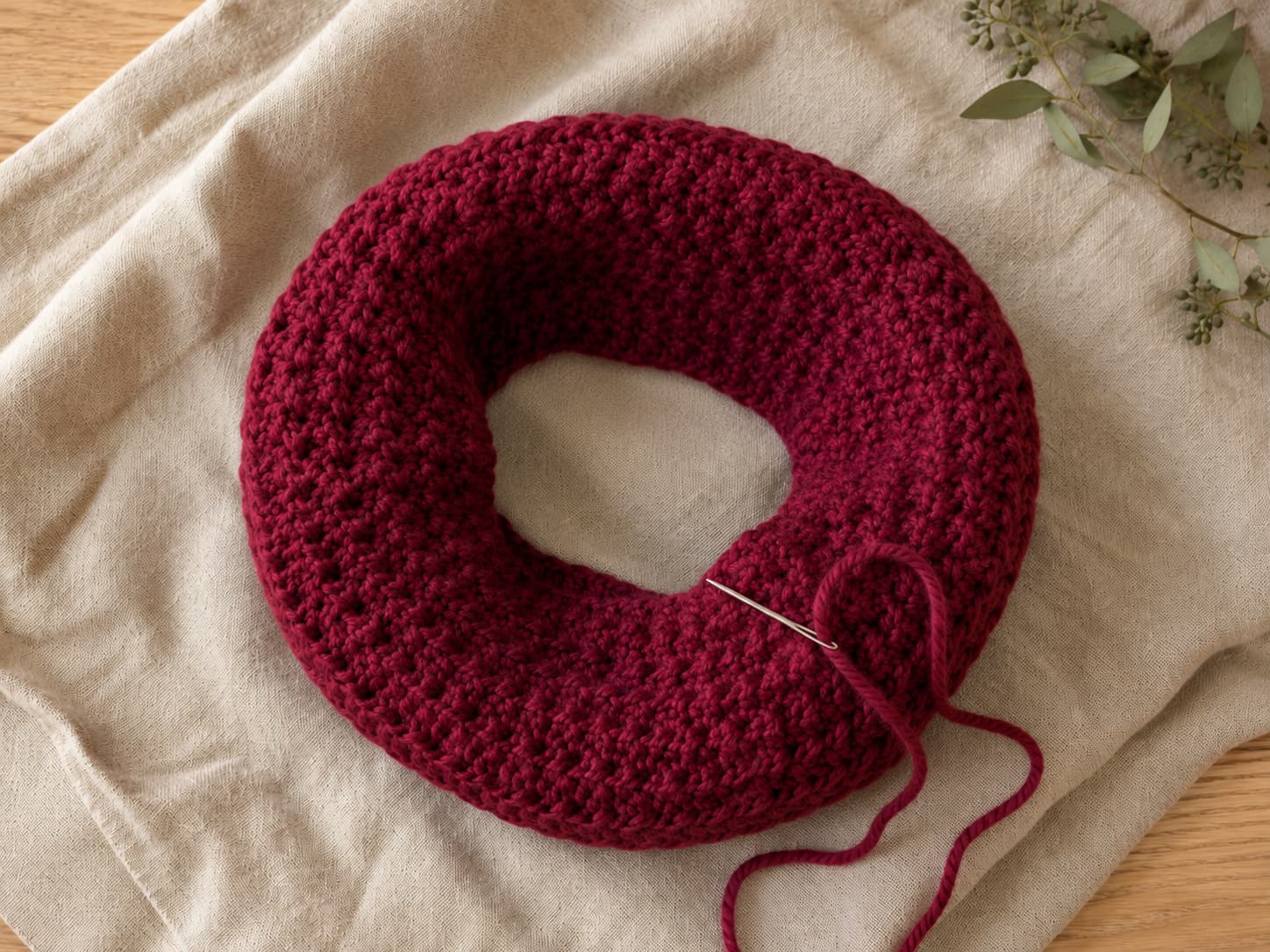 Deep cranberry crochet chunky cowl laid flat on warm linen while a blunt tapestry needle weaves in the final yarn tail near the joined-round seam.