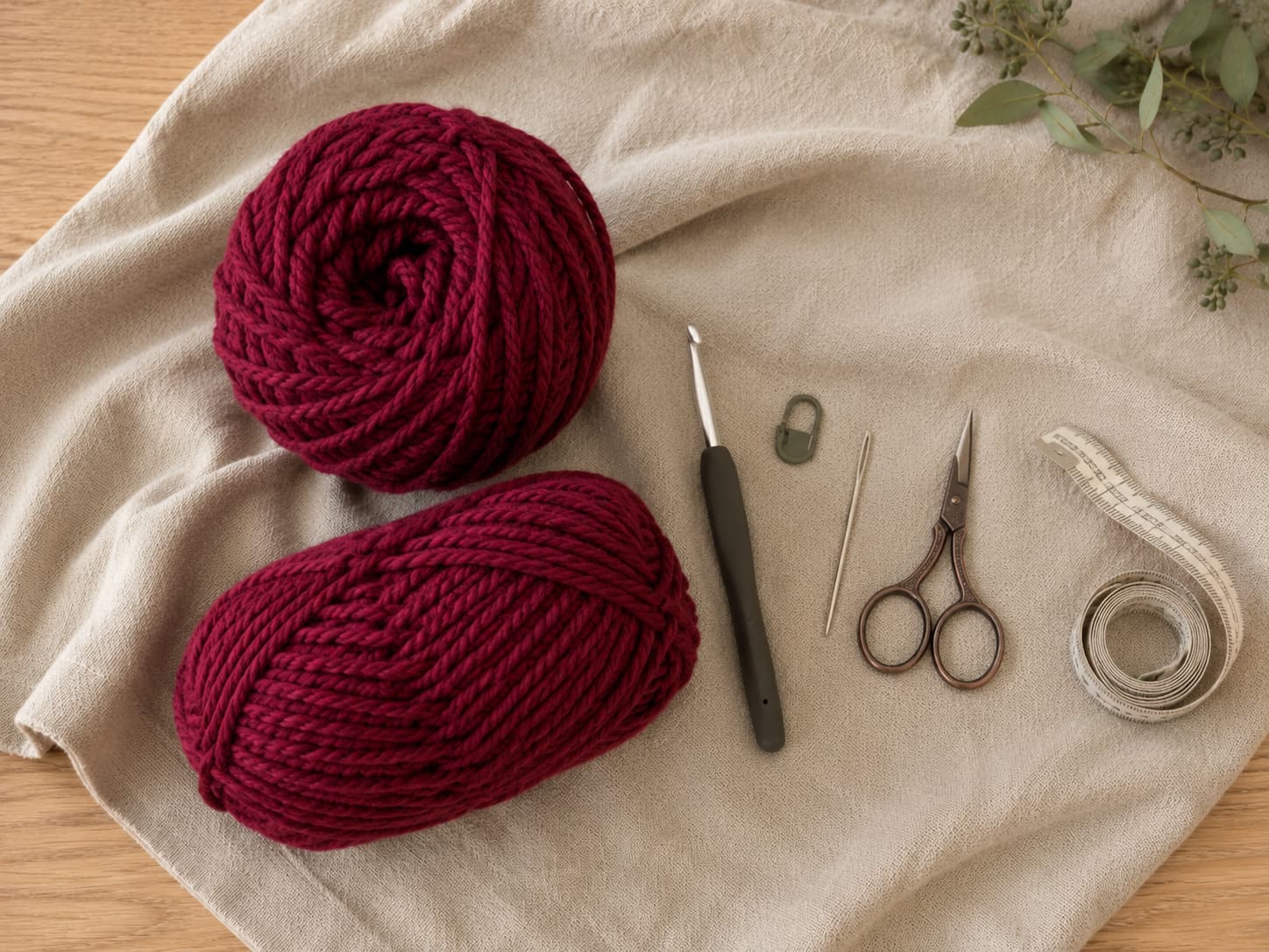 Deep cranberry bulky yarn with a 6.5 mm crochet hook, locking stitch marker, tapestry needle, scissors, and measuring tape on warm linen.