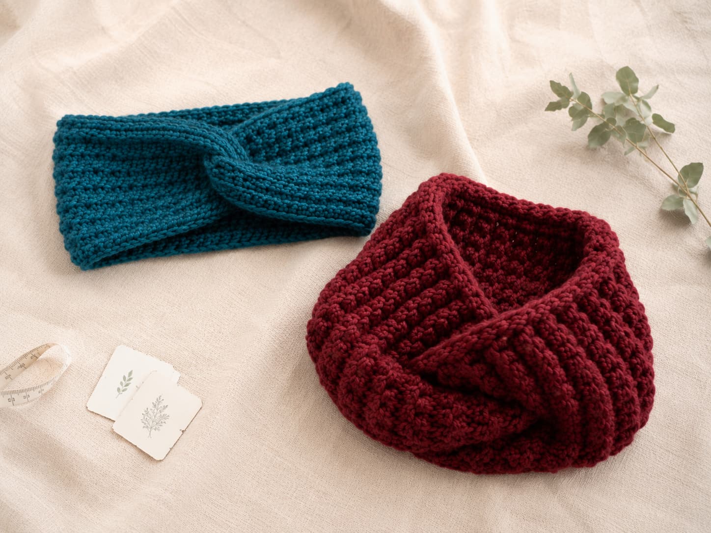 Peacock-teal twisted crochet headband and cranberry chunky cowl folded beside a tape measure and care cards on natural linen.