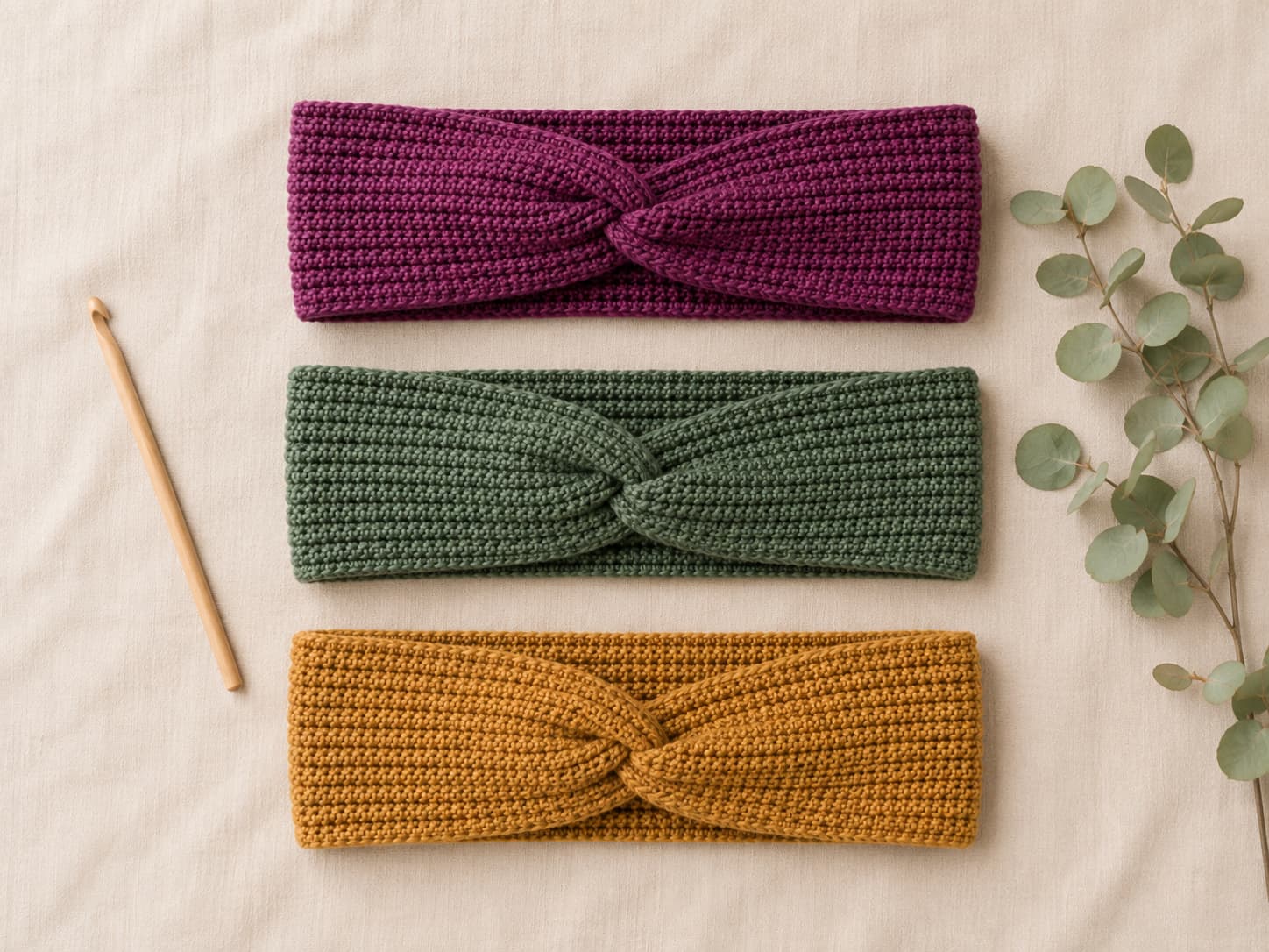 Three finished crochet twisted headbands in soft plum berry, muted forest sage, and golden ochre arranged on warm oatmeal linen with matte wooden accessories.