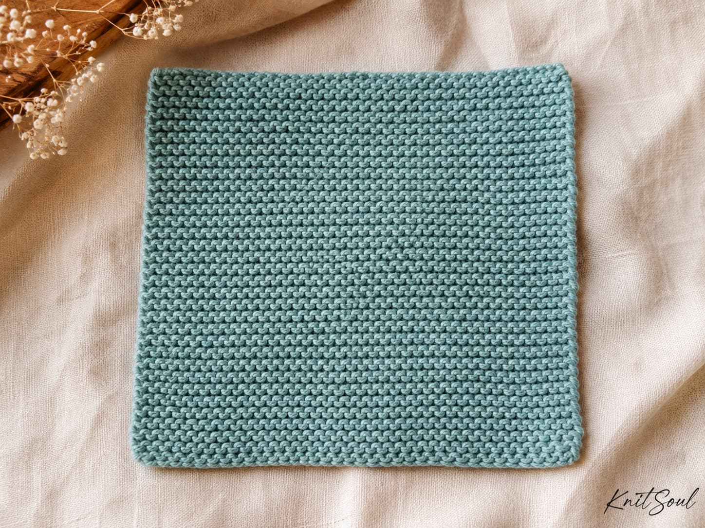 Dusty-aqua square knitted dishcloth worked entirely in garter stitch with even ridges, flexible edges, and slight natural hand-knit variation.