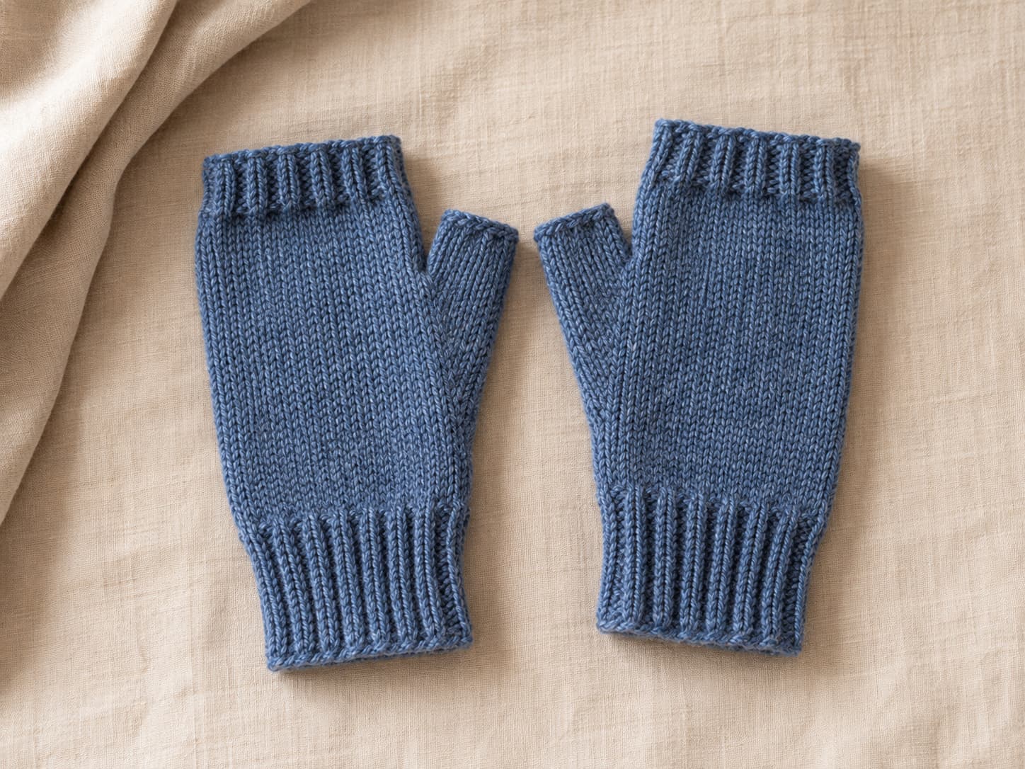 Finished pair of heathered denim-blue knit fingerless mitts with ribbed cuffs and shaped thumb openings arranged on warm natural linen.