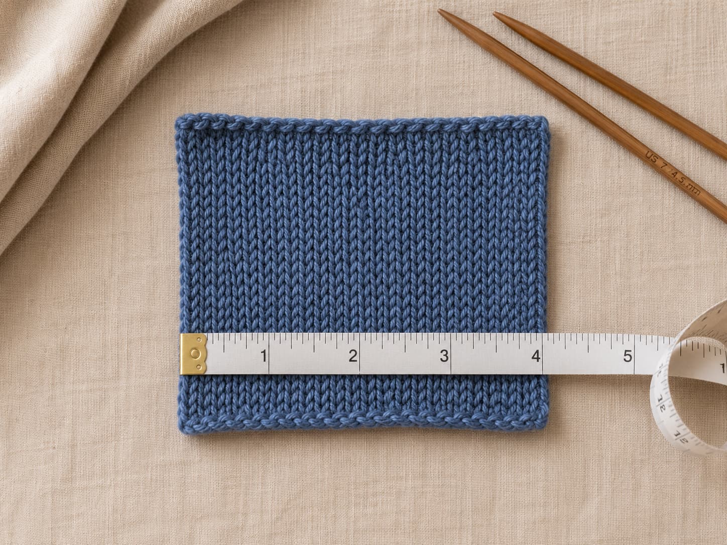 Stockinette gauge swatch in heathered denim worsted yarn measured across four inches beside US 7 knitting needles on warm linen.