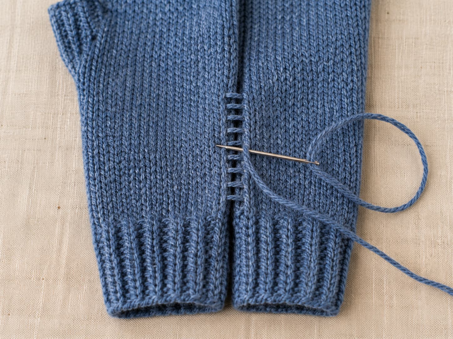 Flat heathered denim fingerless mitt being joined with mattress stitch along the side edge using a blunt tapestry needle on warm linen.