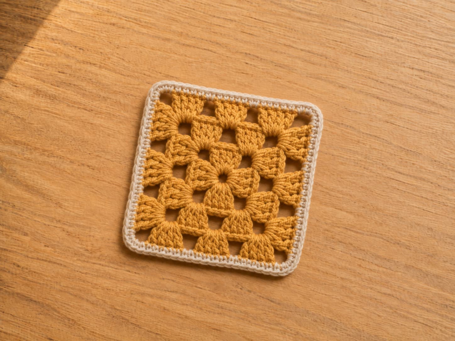 Golden-ochre mini granny square coaster with three rounds of double crochet clusters, clear chain-two corners, and a soft-oatmeal single crochet border.
