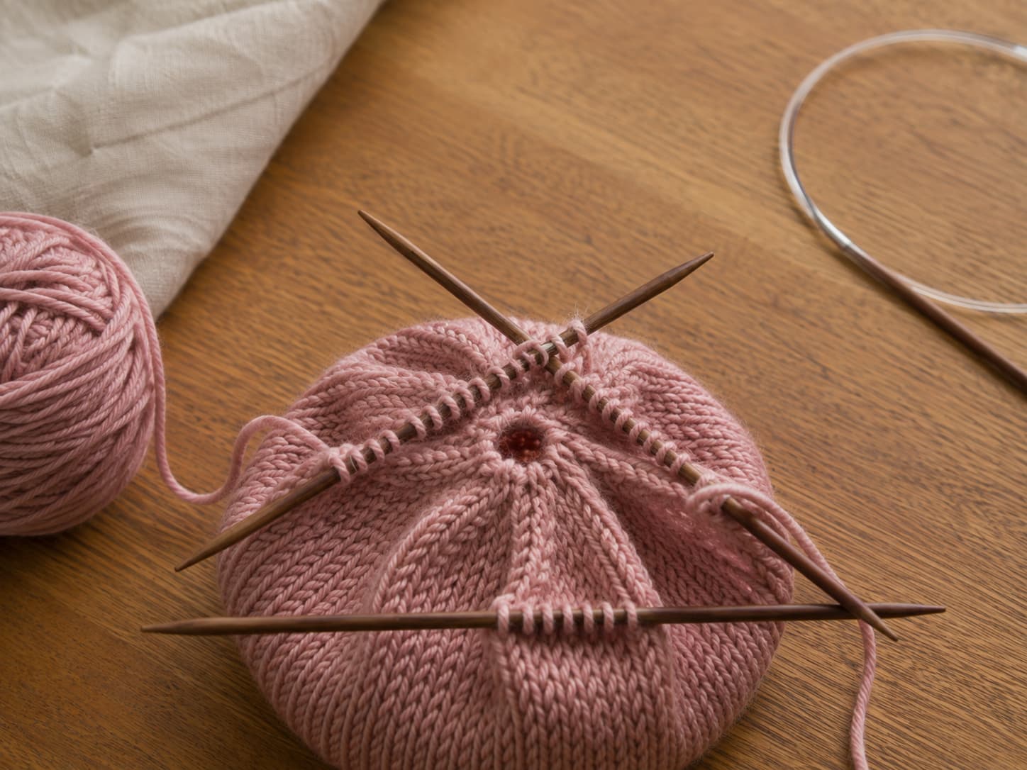 Dusty-rose hat crown with the remaining live stitches distributed across double-pointed needles beside a long circular needle arranged for Magic Loop.