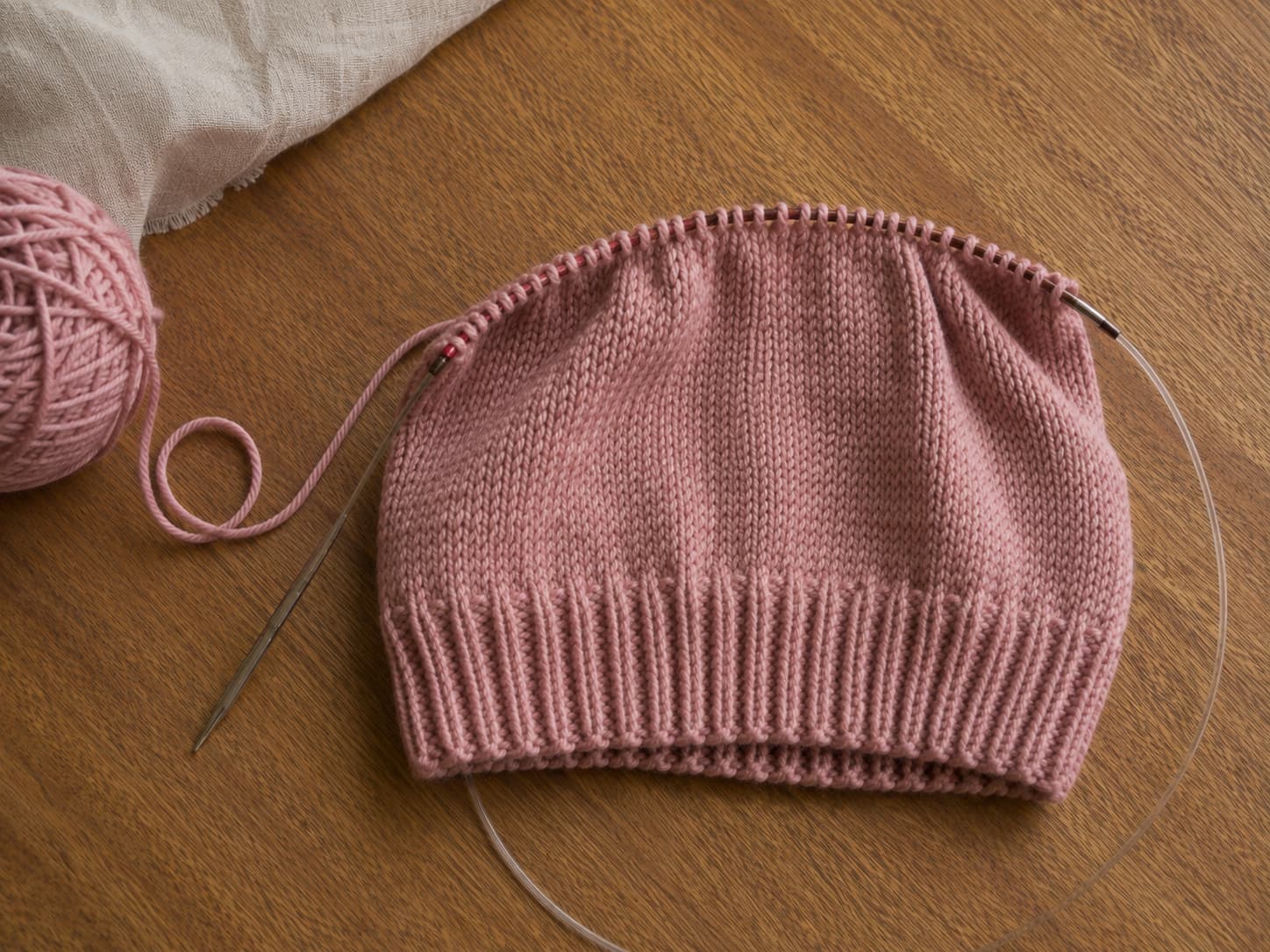 Dusty-rose knit hat in progress showing a single-layer 1×1 ribbed brim transitioning to smooth stockinette on circular needles.