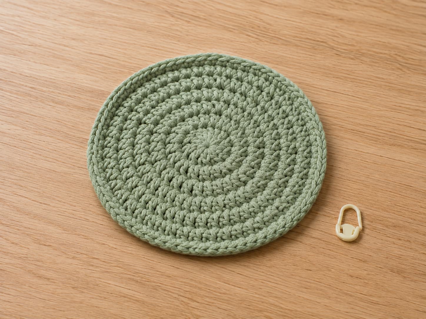 Muted-sage circular crochet coaster with four even half-double crochet increase rounds and a back-loop single crochet border lying flat beside a stitch marker.