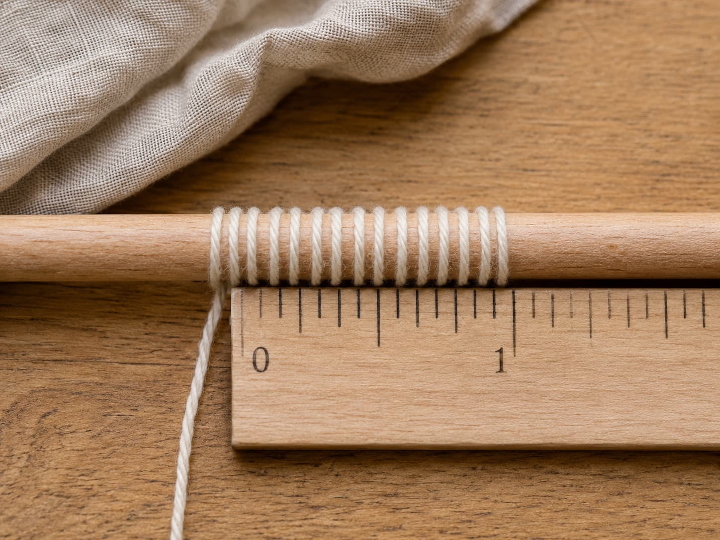 Cream yarn wrapped evenly around a smooth gauge tool beside a ruler to measure wraps per inch without stretching.
