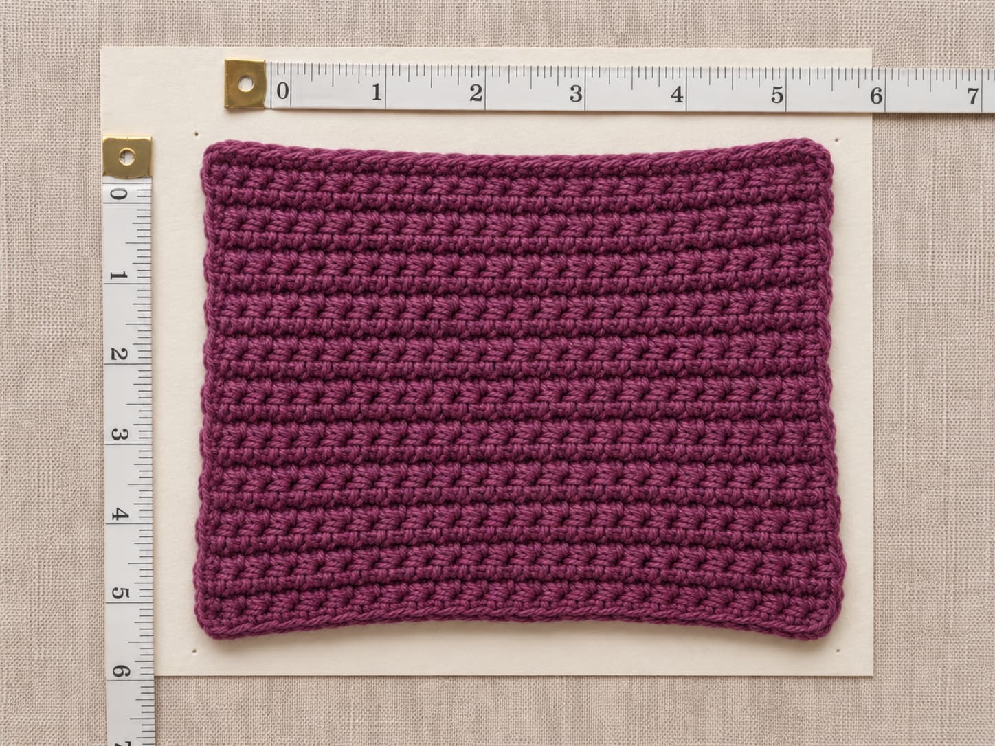 Plum-berry half-double crochet swatch beside a tape measure showing fourteen stitches and eleven rows across a four-inch square on warm off-white paper.