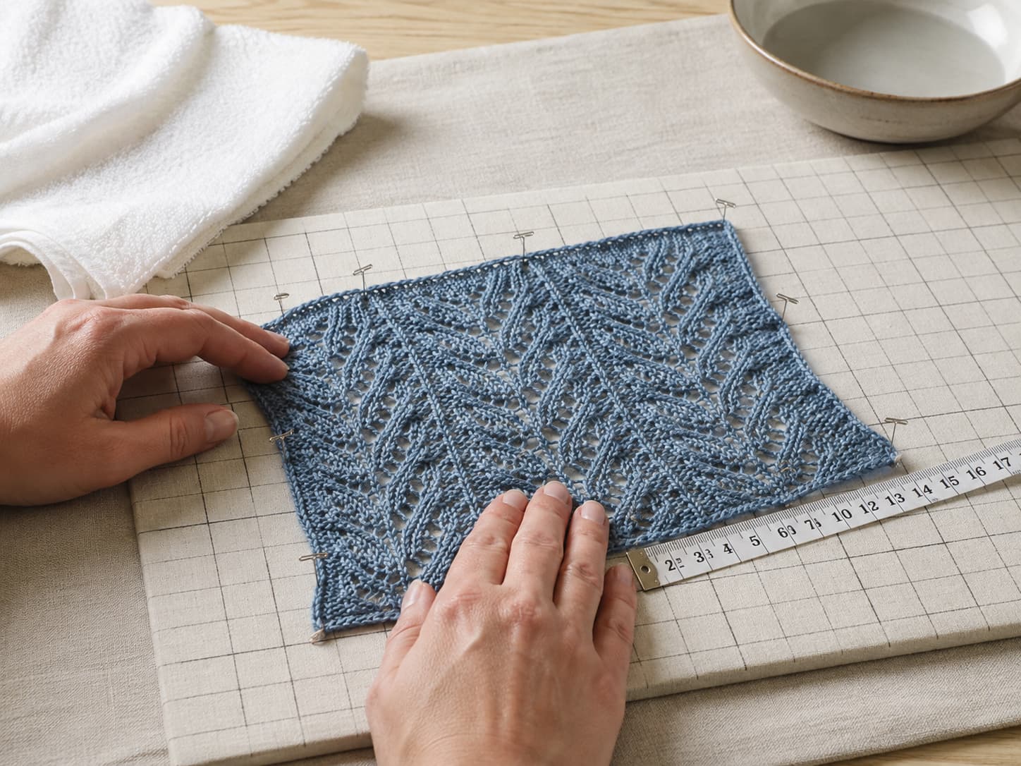 Mineral-blue lace swatch being supported in water, rolled in a towel, measured, and pinned gently to shape on a gridded blocking mat.