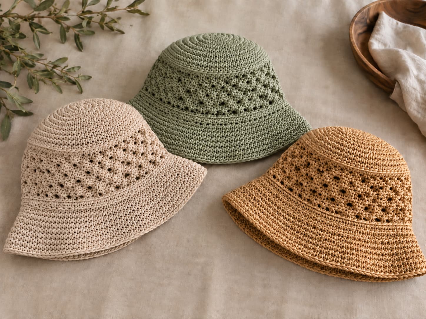 Three crochet bucket hat variations in oatmeal, sage, and raffia-style yarn on a warm neutral background