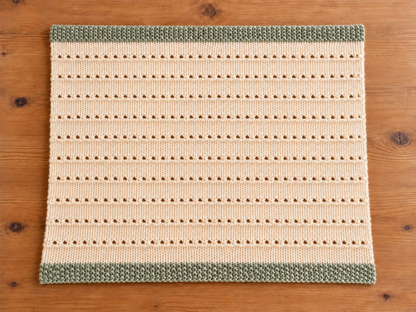Finished cream knit baby blanket with evenly spaced horizontal Cloud Nine eyelet rows and pale sage seed stitch borders on a natural wood surface.