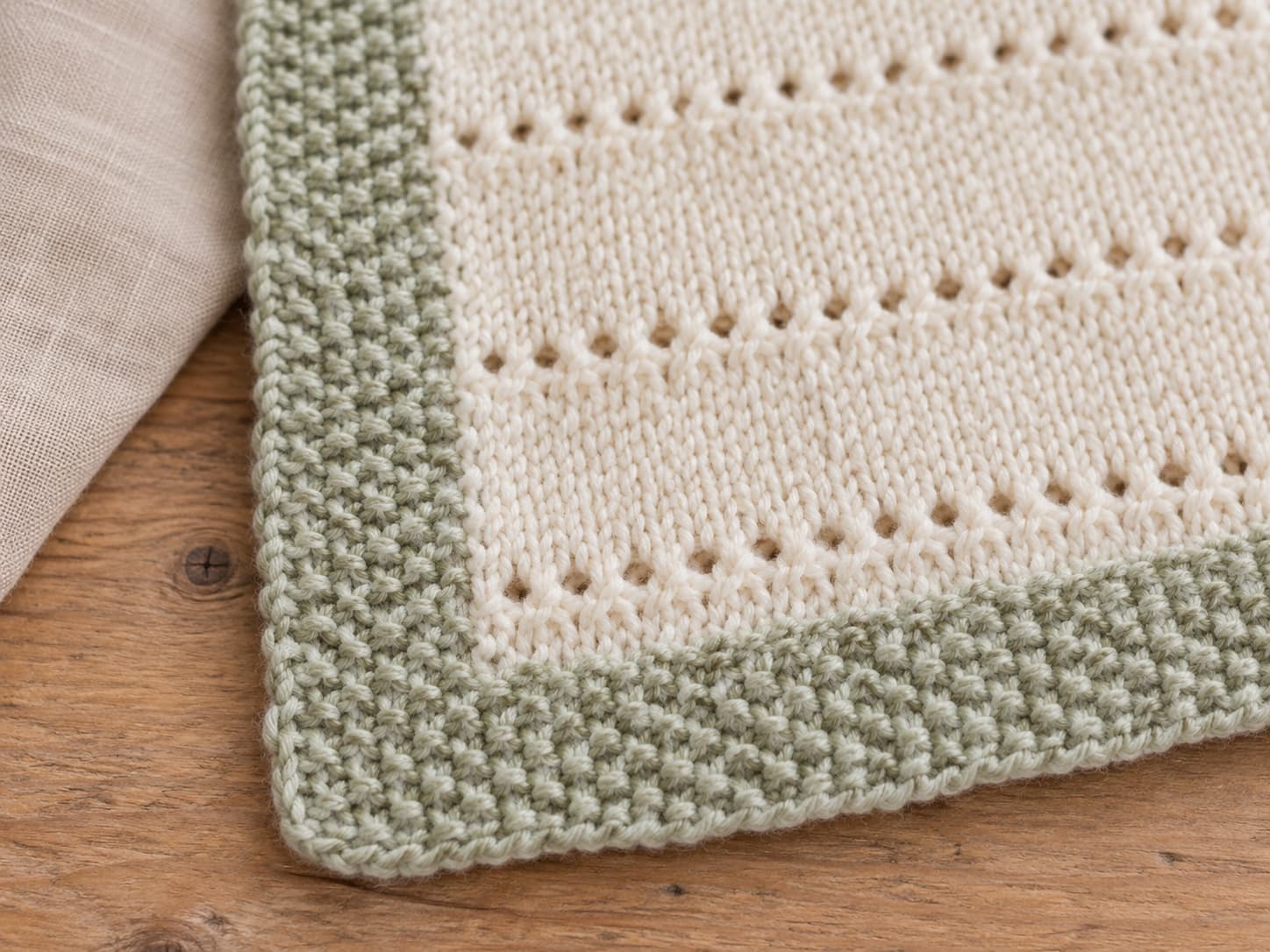 Pale sage seed stitch border at the edge of a cream Cloud Nine Stitch knit baby blanket showing the bumpy alternating texture.